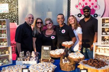 Group of people smiling and posing behind table laden with a variety of small cupcakes