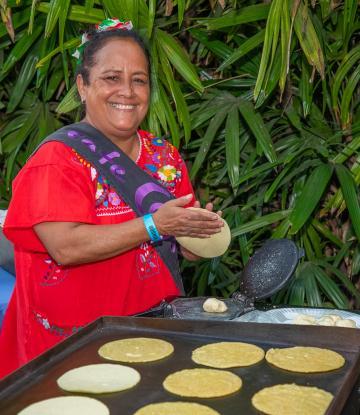 Woman smiling and shaping tortillas being placed on a large griddle 